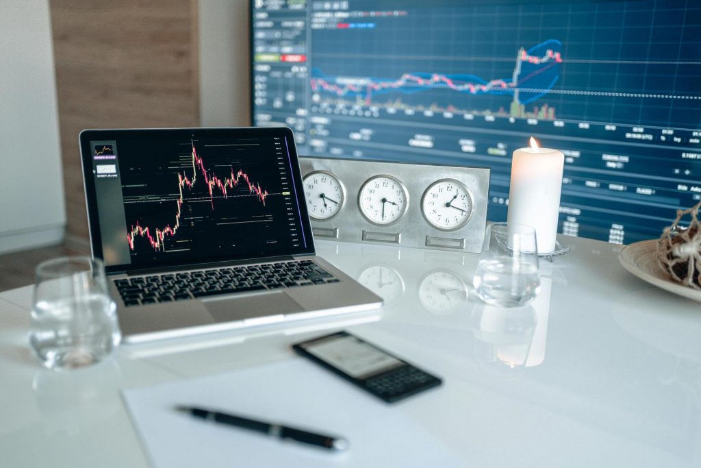 Mesa de trabajo con un portátil mostrando gráficos de trading, una pantalla con cotizaciones bursátiles, relojes de zonas horarias y una vela encendida, representando análisis financiero.
