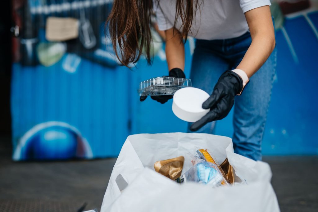 Persona con guantes recogiendo residuos plásticos y depositándolos en una bolsa, como acción de limpieza y reciclaje urbano.