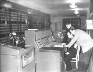 Ingenieros trabajando con un gran ordenador antiguo en sala llena de paneles electrónicos