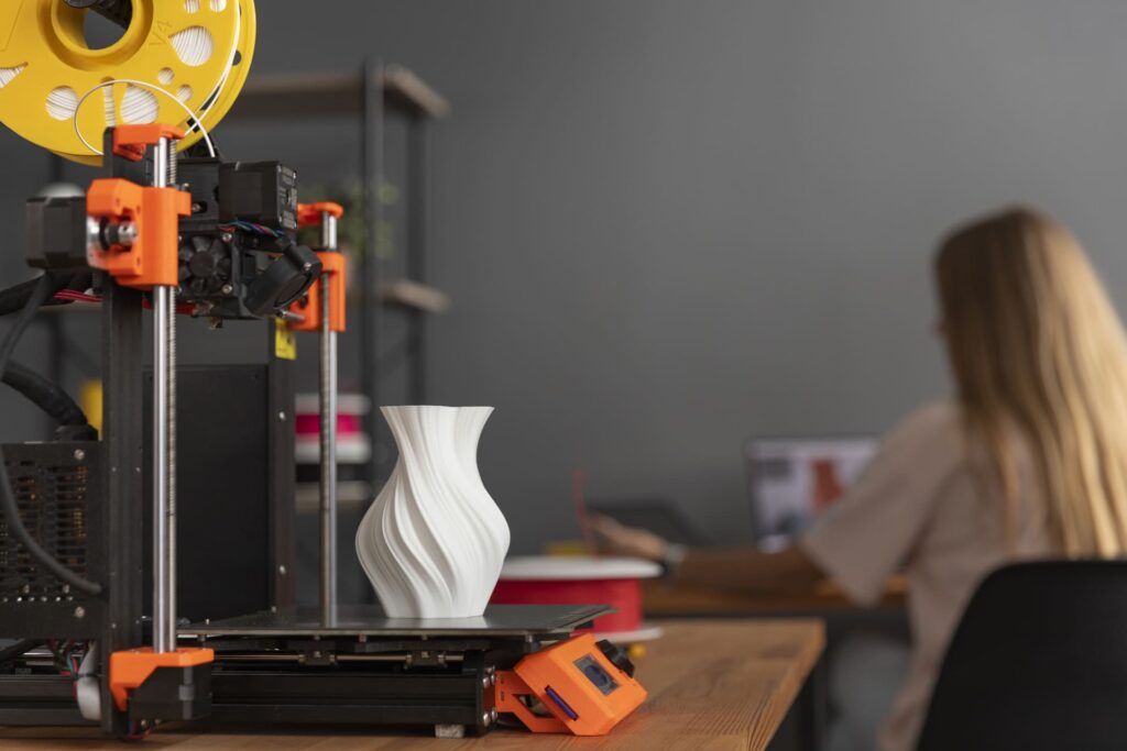 Impresora 3D fabricando un jarrón blanco sobre una mesa de trabajo, con una persona diseñando en un ordenador al fondo.