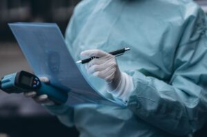 Persona con equipo de protección sanitaria y guantes revisando un documento mientras sostiene un dispositivo de medición en un entorno clínico o industrial.