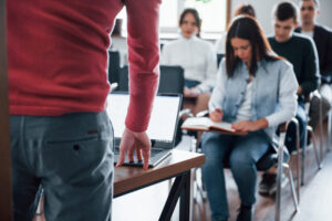 Profesor impartiendo una clase presencial mientras varios alumnos toman apuntes en un aula moderna con un portátil sobre la mesa.