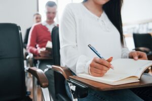 Primer plano de una persona tomando apuntes en una libreta durante una clase o conferencia, con varios asistentes desenfocados al fondo en un aula moderna.